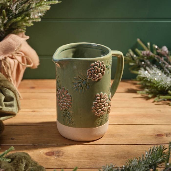 Festive stoneware jug with deep green glazed upper body, natural base, embossed pinecones and pine branches, and single handle